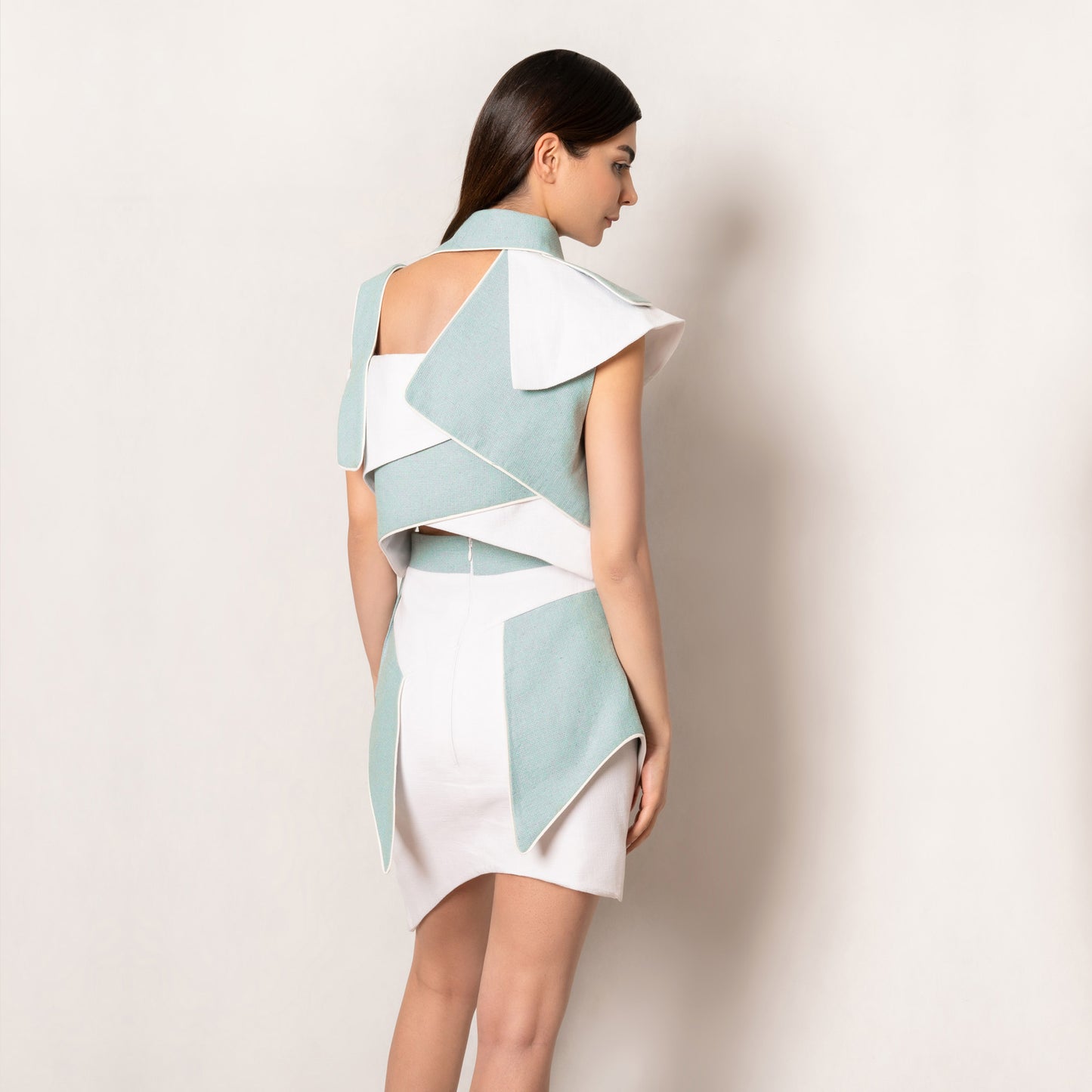 The back showcases intersecting panels that create a criss-cross architectural finish. A sharp cut-out at the upper back adds edge, while the skirt’s structured mint detailing frames the silhouette with precision.