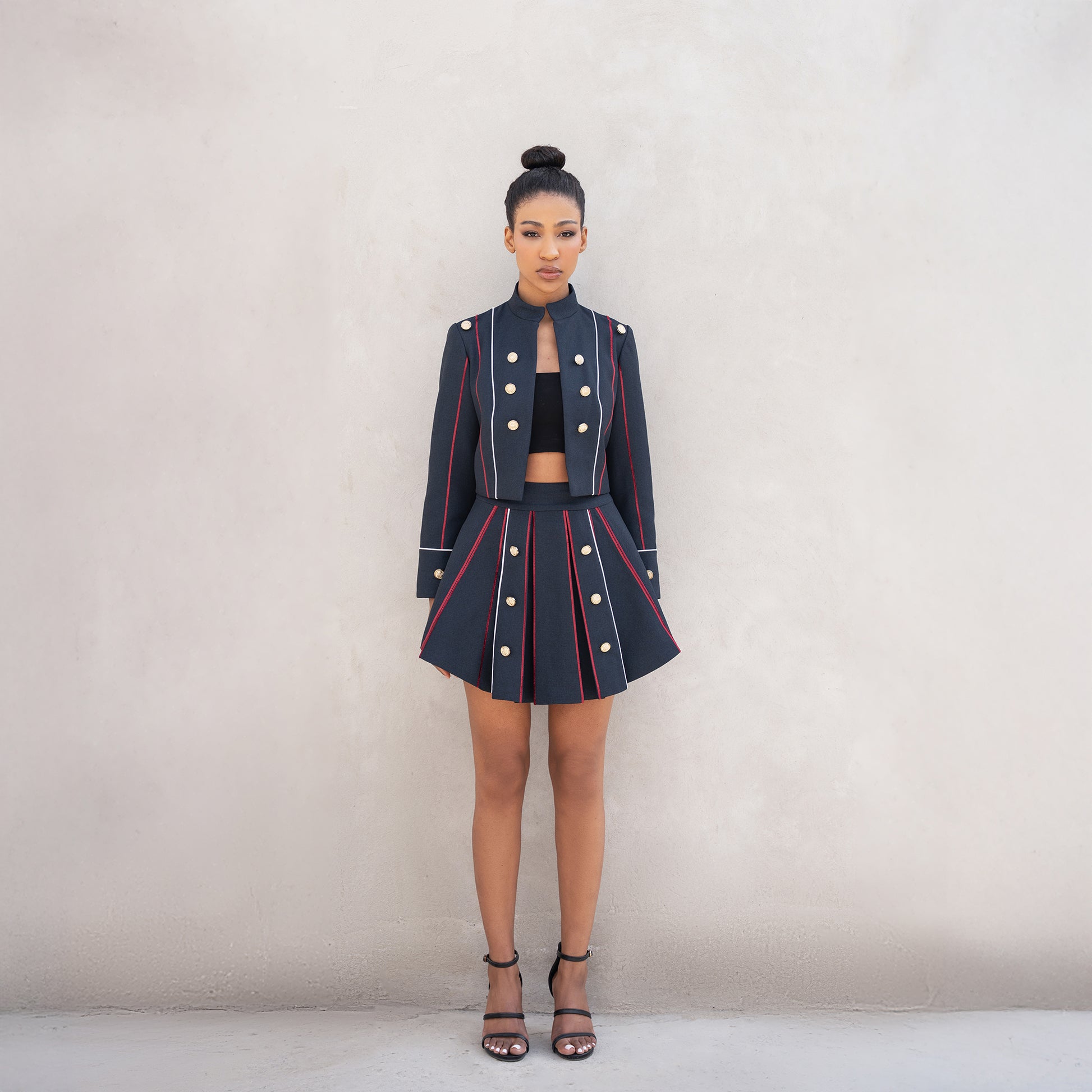 A woman wearing the Axis set, consisting of a cropped Fastoni jacket with golden buttons and a pleated skirt with red and white laser-cut lines.