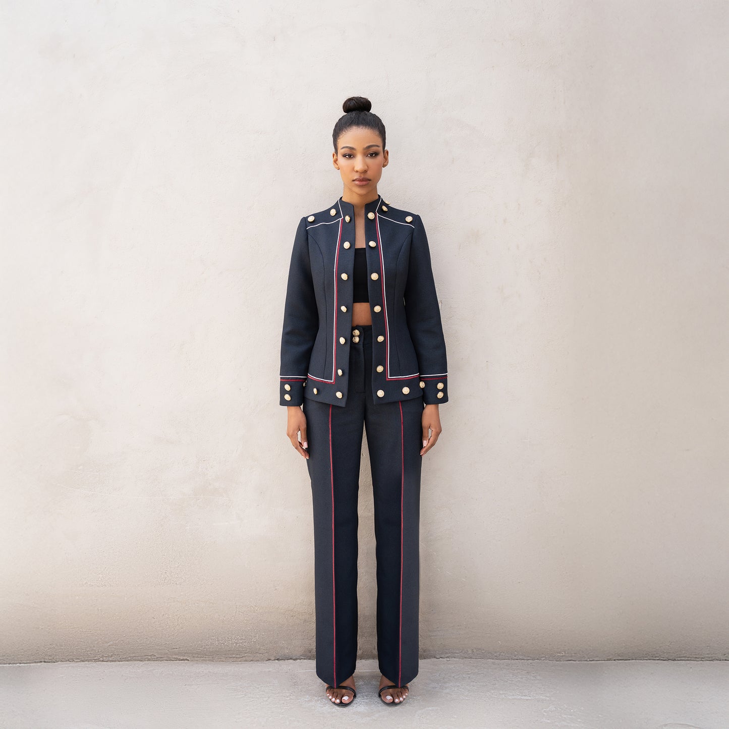 A woman wearing the Frame suit, tailored from black Fastoni fabric with red and white laser-cut lines and golden buttons on the jacket and trousers.